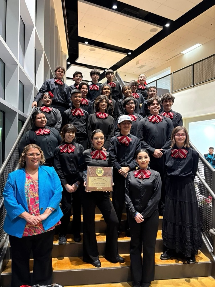 Mariachi students in black uniforms with red bow ties pose on a staircase holding a championship plaque, standing beside their director.