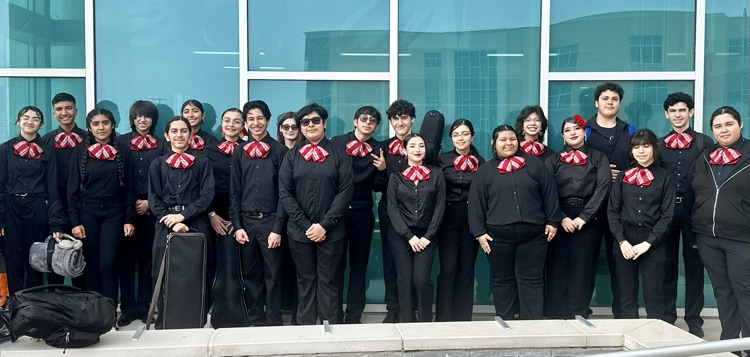Mariachi students in black uniforms with red bow ties pose on a staircase holding a championship plaque, standing beside their director.