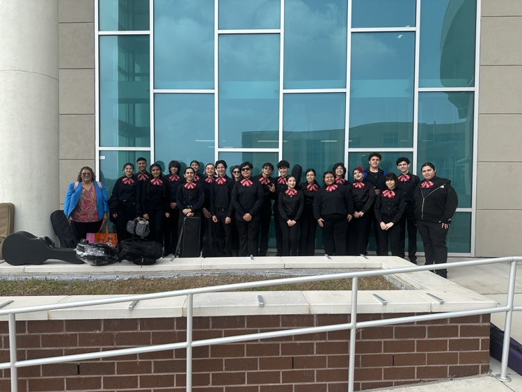 Mariachi students in black uniforms with red bow ties pose on a staircase holding a championship plaque, standing beside their director.
