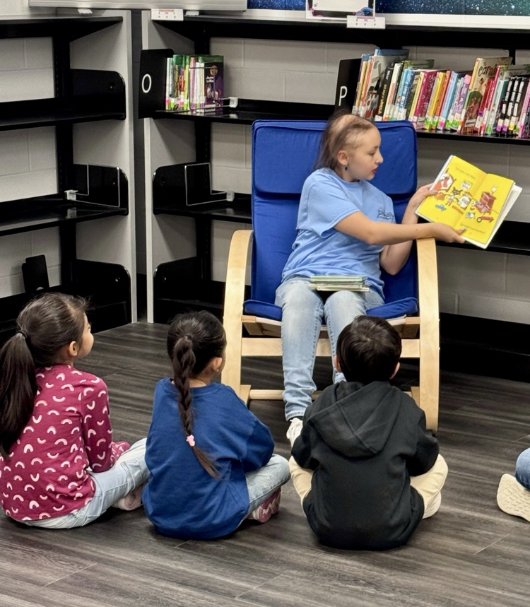 Alt text: High school students read picture books to elementary children in a colorful school library setting.