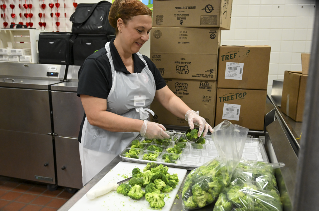 A woman in a black shirt and white apron prepares broccoli in a commercial kitchen.