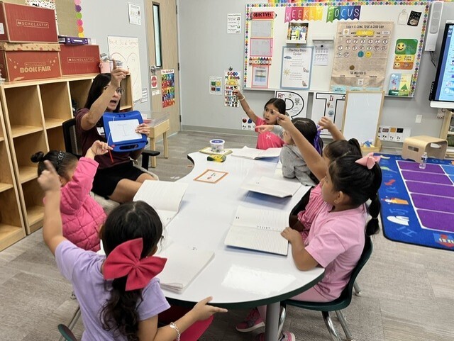 A woman and four girls seated at a table in a classroom, raising their hands while holding tablets.