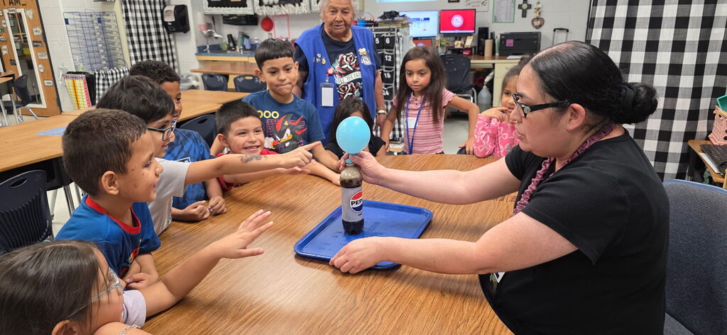 A teacher holds a balloon and a tray in a classroom; children gather around and watch.