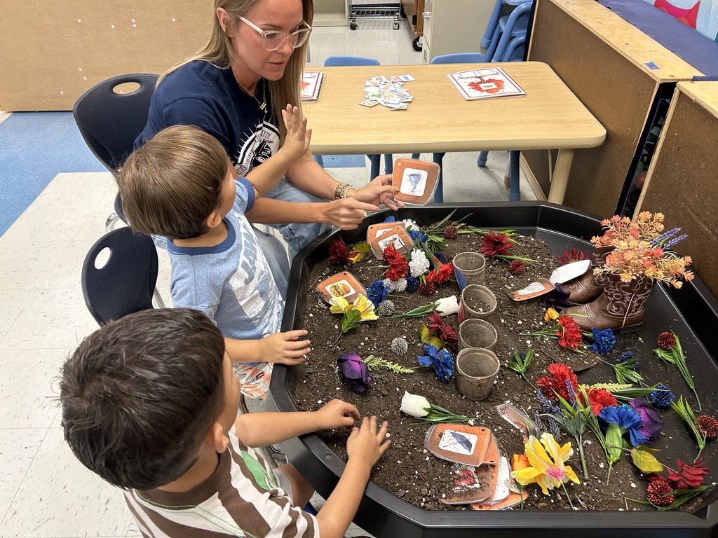 Woman and two young children seated around a table with soil and plastic flowers. Table has small flowers and plants.