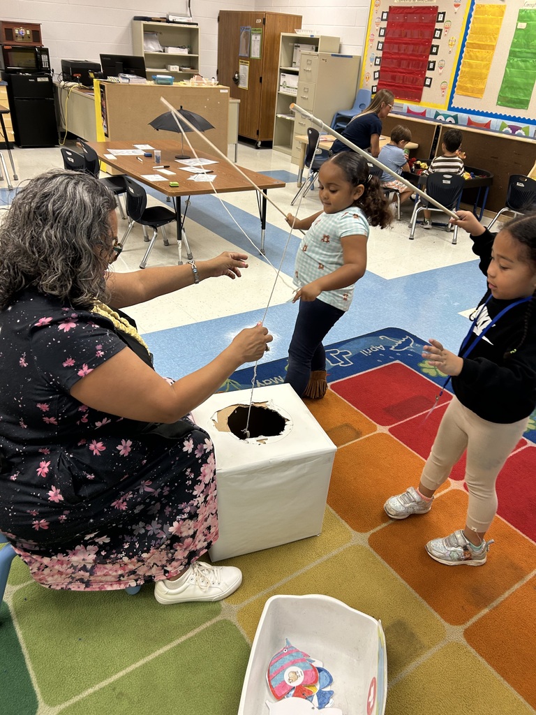 An adult and two children pull on a rope inside a classroom. A colorful rug covers the floor.