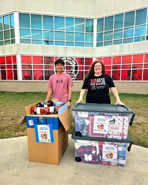 Two people standing next to boxes of donated items, with a large building, Baker Middle School, in the background.