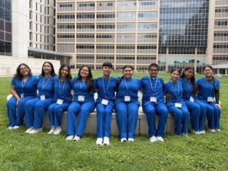 A group of people in blue uniforms sit on a bench, with buildings in the background.