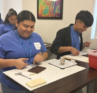 Two people in blue shirts work on a project at a table. One uses scissors, and the other holds a small object.