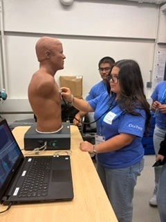 A person uses a stethoscope on a mannequin, while another individual checks a laptop. Two others watch.