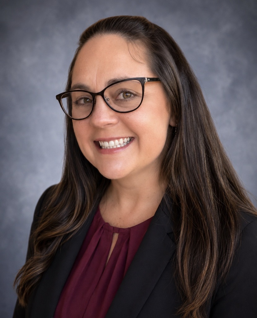 A smiling woman with long brown hair wearing glasses and a black jacket, standing in front of a gray background.