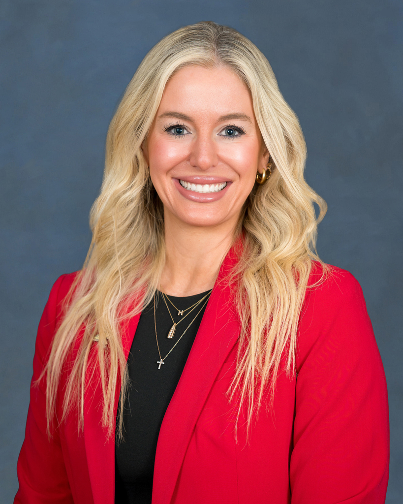 Woman with long blonde hair wearing a red blazer and a black top. She is smiling and standing against a gray background.
