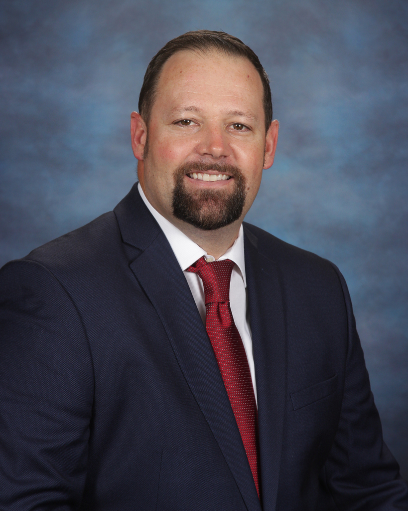 Person in navy suit and red tie, smiling. Background is blue with gradient. Shoulders and upper chest are visible.