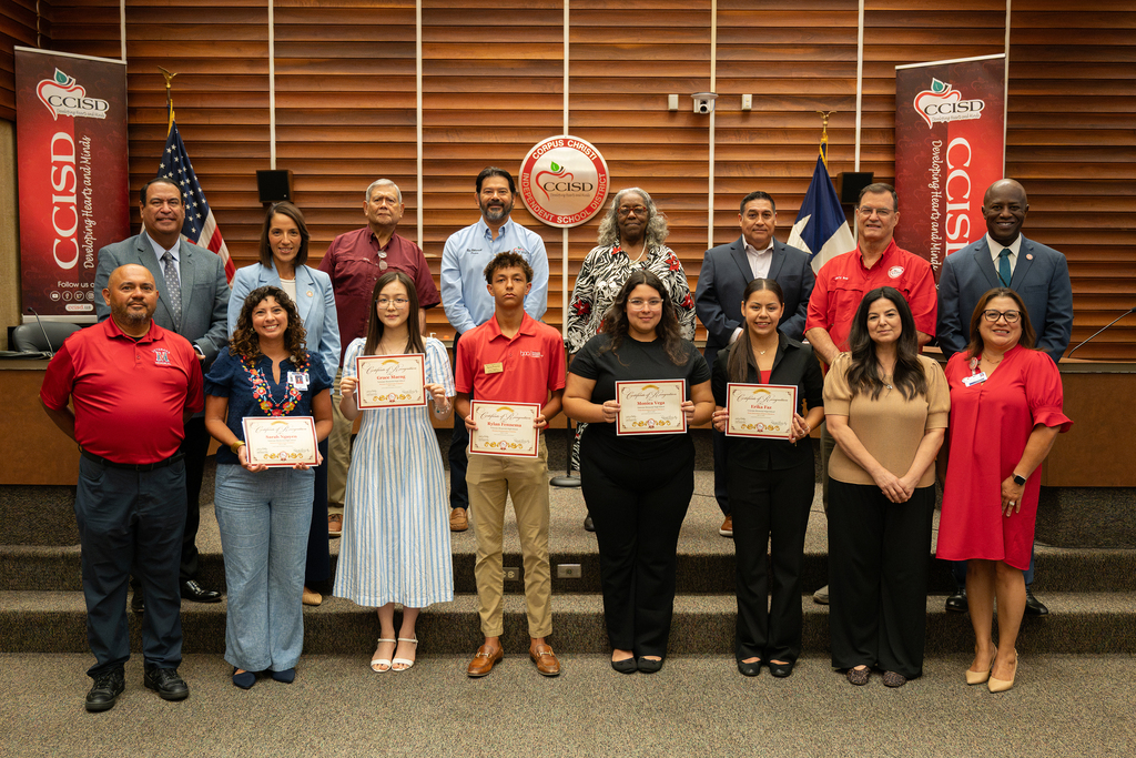 Group of people in a room posing for a photo. Some hold certificates. Flags in the background.