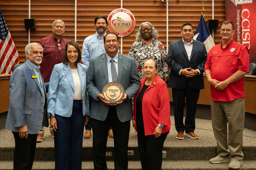 A group of people standing on stairs, one person holds an award. A red sign with "CCISD" and flags are behind them.