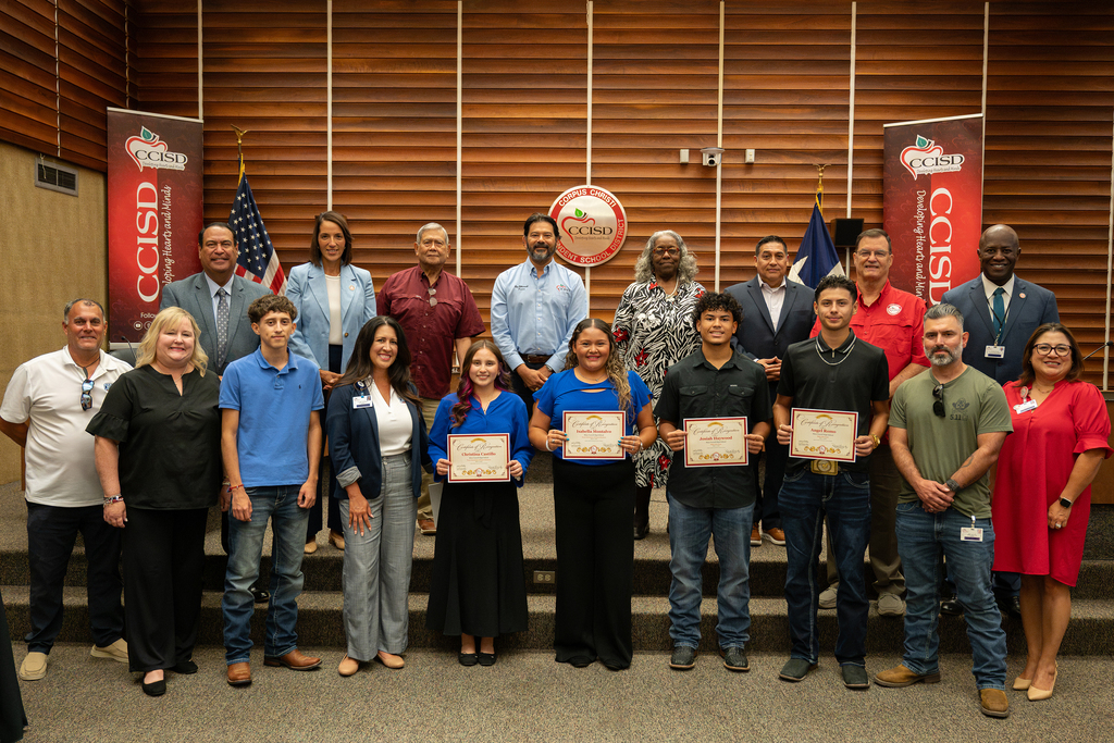 A group of people in business attire stands on a stage holding certificates. Behind them are banners and flags.