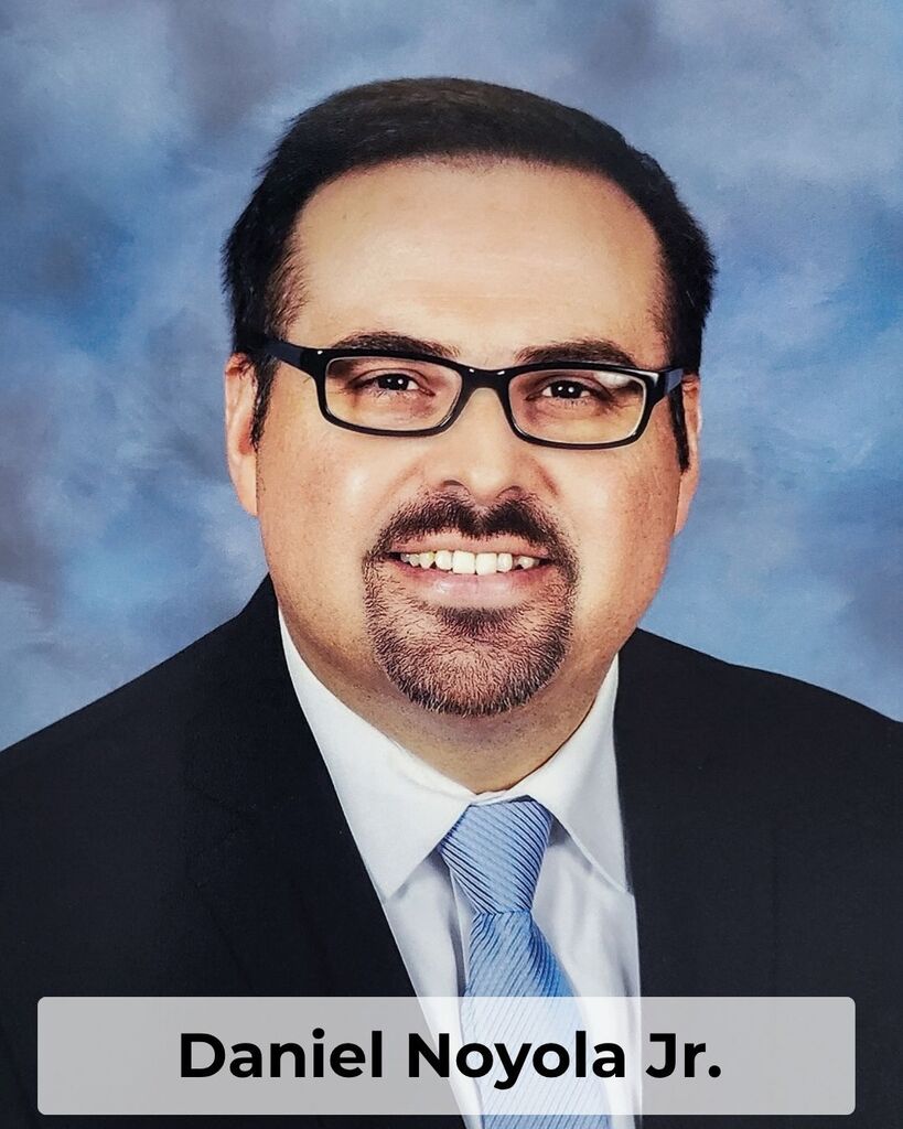 A smiling man in a suit, tie, and glasses poses for a photo against a blue background. The text reads "Daniel Noyola Jr."