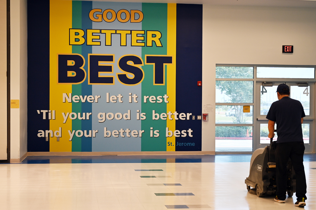 A person with a push broom cleaning the floor, with a wall mural behind that reads "Good Better Best."