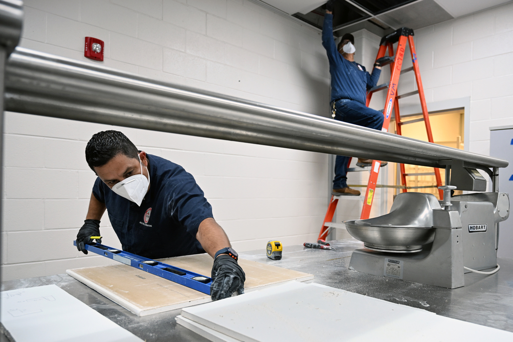 Two people are working in a room. One is standing on a ladder and another is measuring.