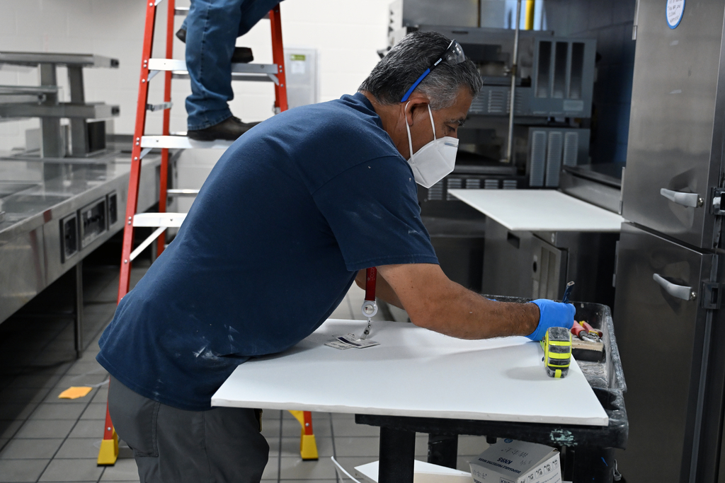 Man in blue shirt and face mask, wearing gloves, works on a table in a commercial kitchen.
