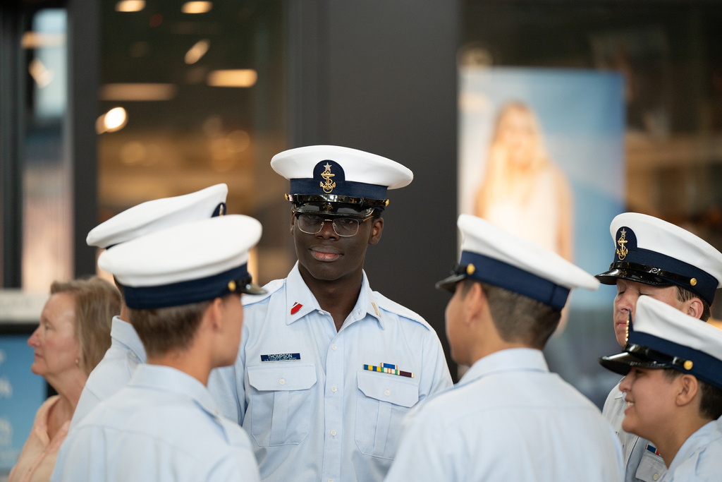 Group of people in navy uniforms standing in a row, facing left. A man in glasses and a hat stands in the center.