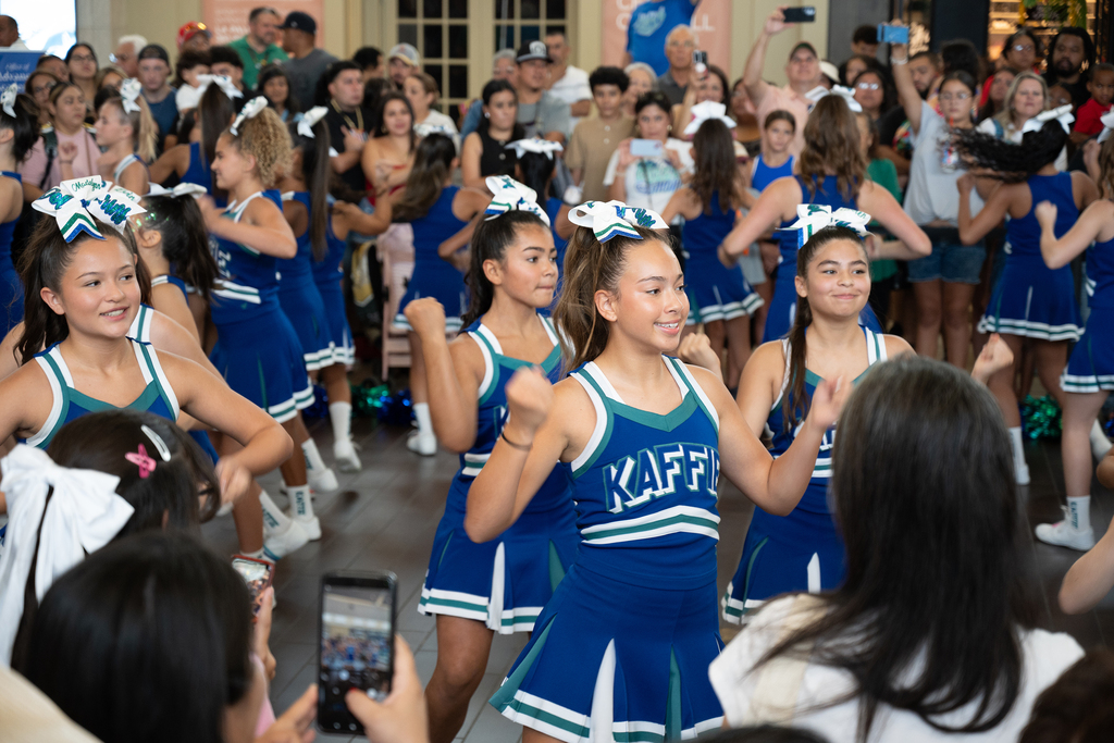 Cheerleaders in blue uniforms with white trims perform in front of an audience. Some are raising their hands.
