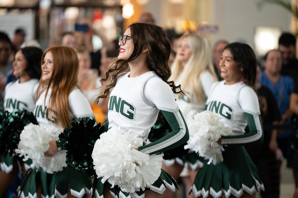 Cheerleaders with white and green uniforms perform in front of an audience in a mall.