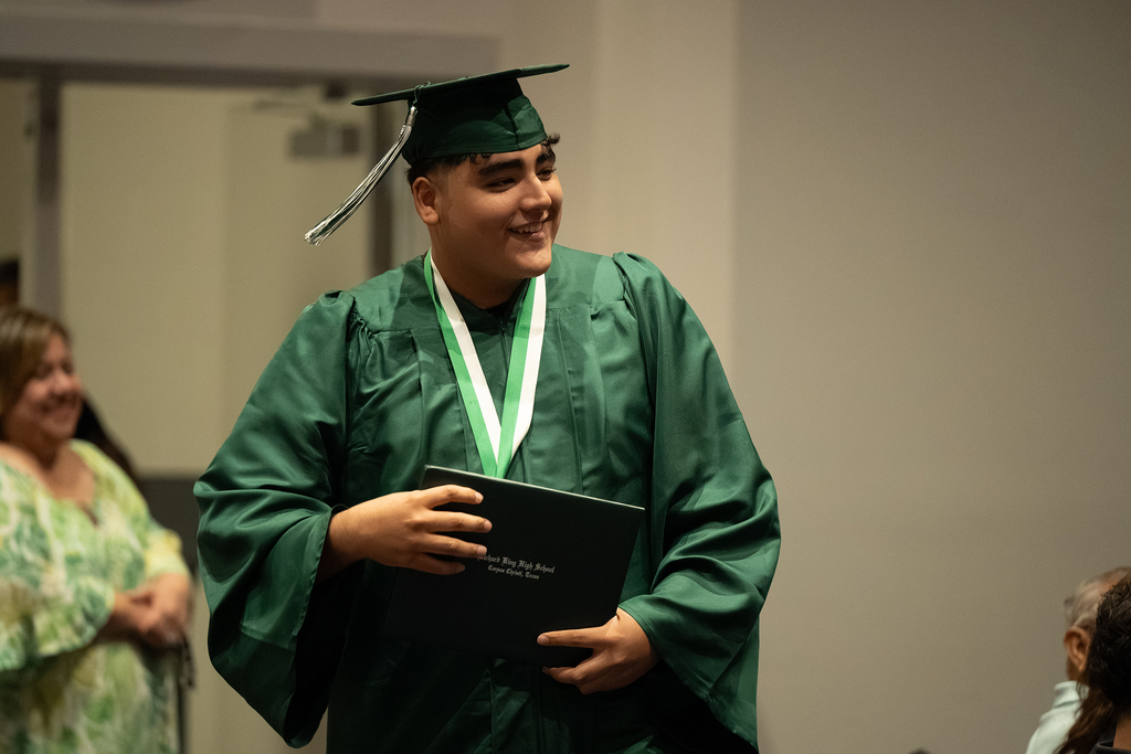 A man in a green graduation gown, hat, and holding a diploma smiles at the camera. Behind him, a woman in a green dress watches.