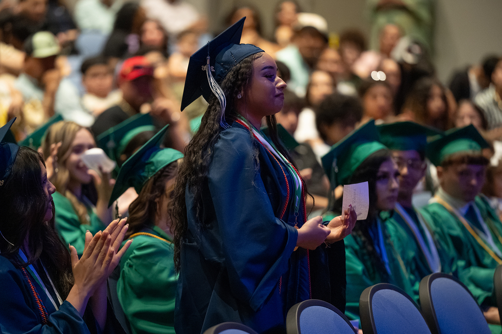A group of people in green gowns and caps are attending an event; one person is standing and clapping.