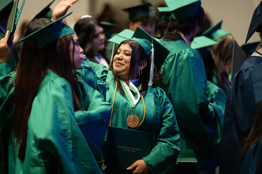 A group of individuals in green graduation gowns and caps stand close together. One person holds a diploma and a medal.