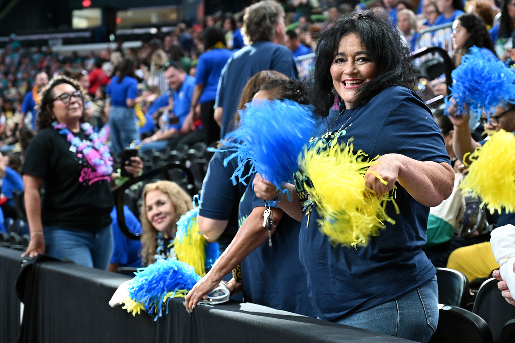 A group of women, one holding blue and yellow pom-poms, are seated in a stadium. Behind them, a crowd is watching.