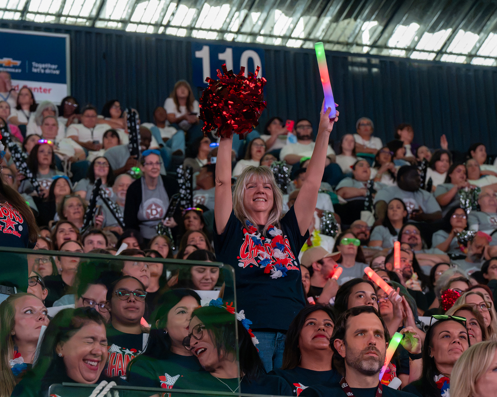 Spectators in a stadium with red and blue decorations, cheering. A woman holds a red pom-pom and a light stick.