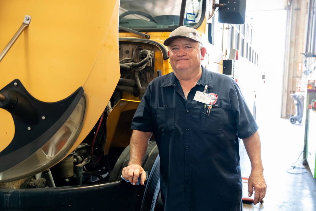A person in a blue shirt and cap stands beside a yellow vehicle in a workshop.