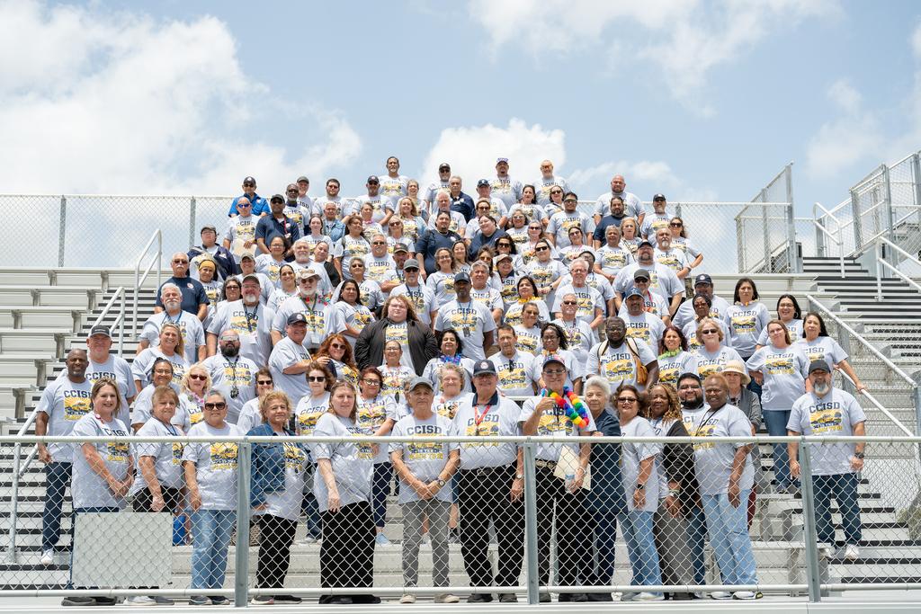 A large group of adults wearing matching white shirts poses for a photo in a stadium.