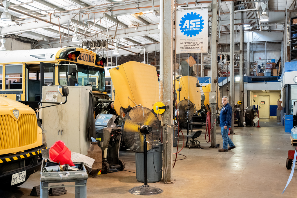 Interior of a garage with several buses parked and a person standing near the vehicles, a blue banner is attached to a pole.