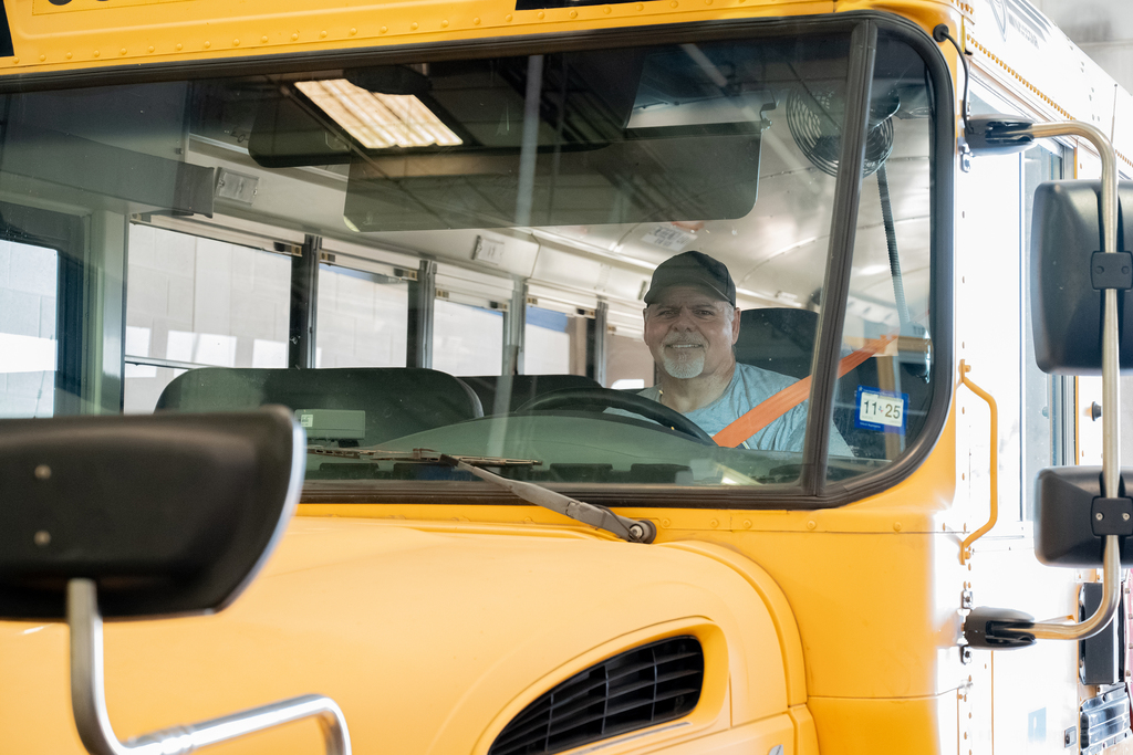 A man seated in a school bus wearing a cap and a seatbelt. The bus is yellow.