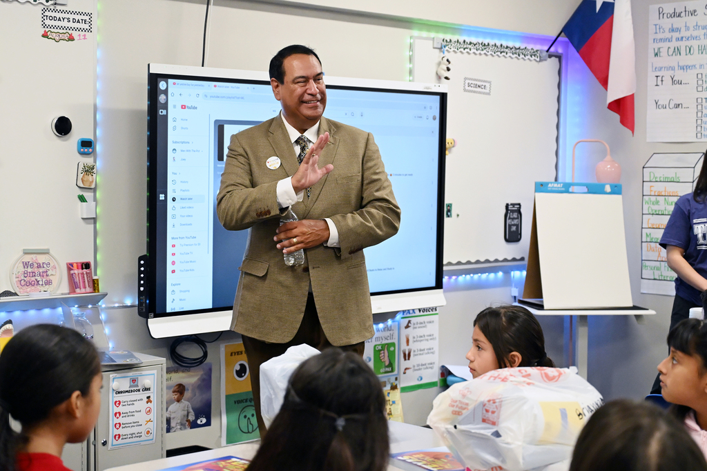 Man in suit stands in front of a screen, presenting to a classroom of children.