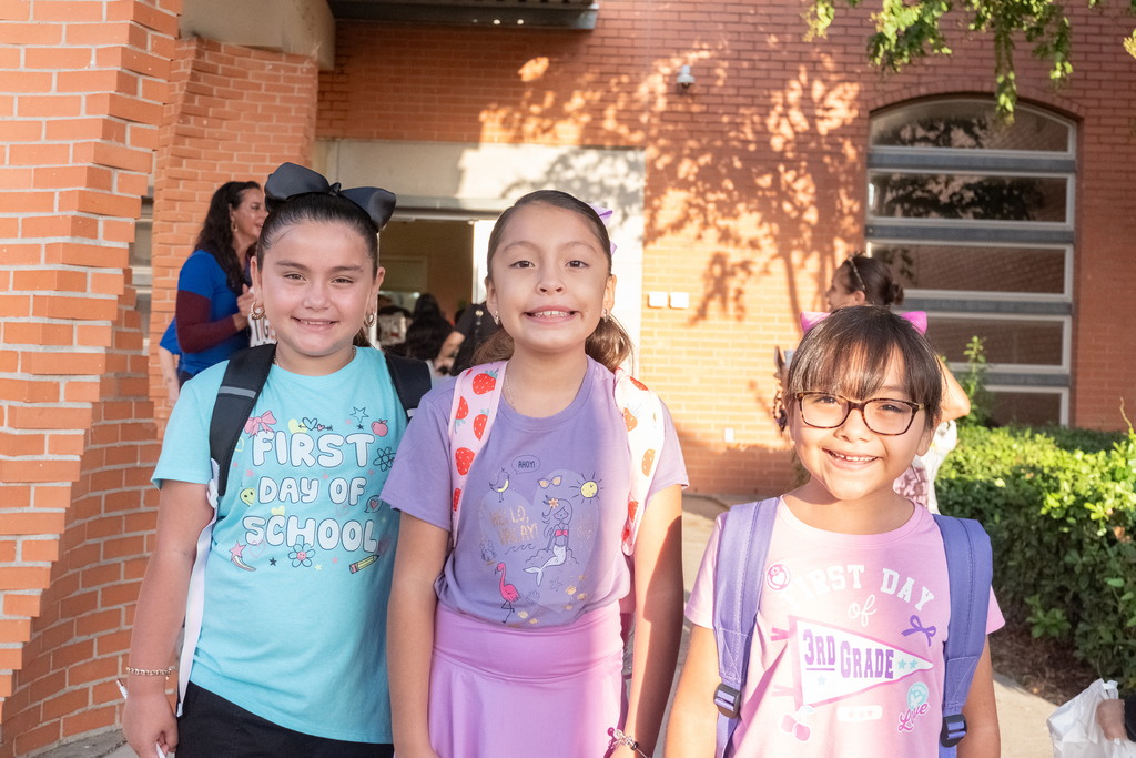 Three girls standing outside a brick building, two with backpacks and one with glasses, smiling.