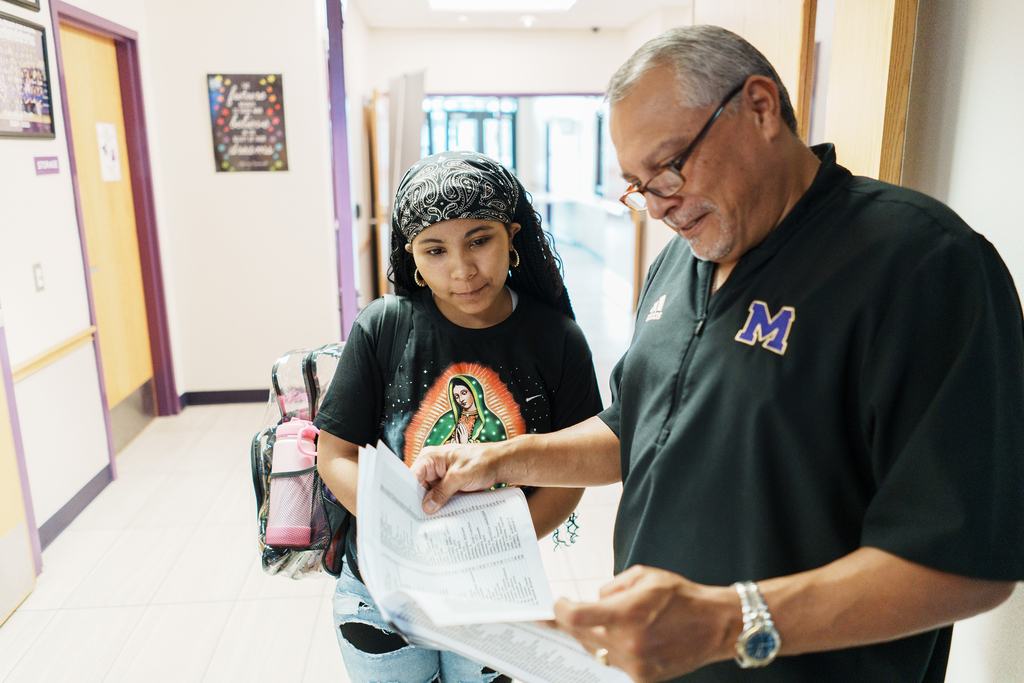 A man and a girl review papers in a hallway. The man wears glasses, a watch, and a black shirt.