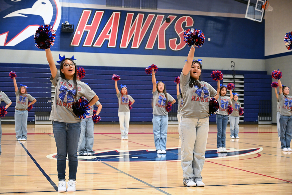 A group of cheerleaders performs on a basketball court, with the word "HAWKS" on a blue wall.