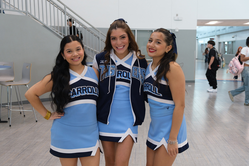 Three cheerleaders in matching blue uniforms pose together in a school hallway with a staircase in the background.