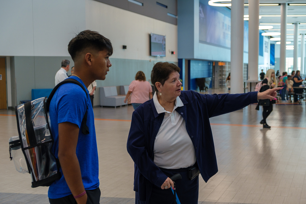 A woman in a navy coat directs a young man in a blue shirt in a modern building.