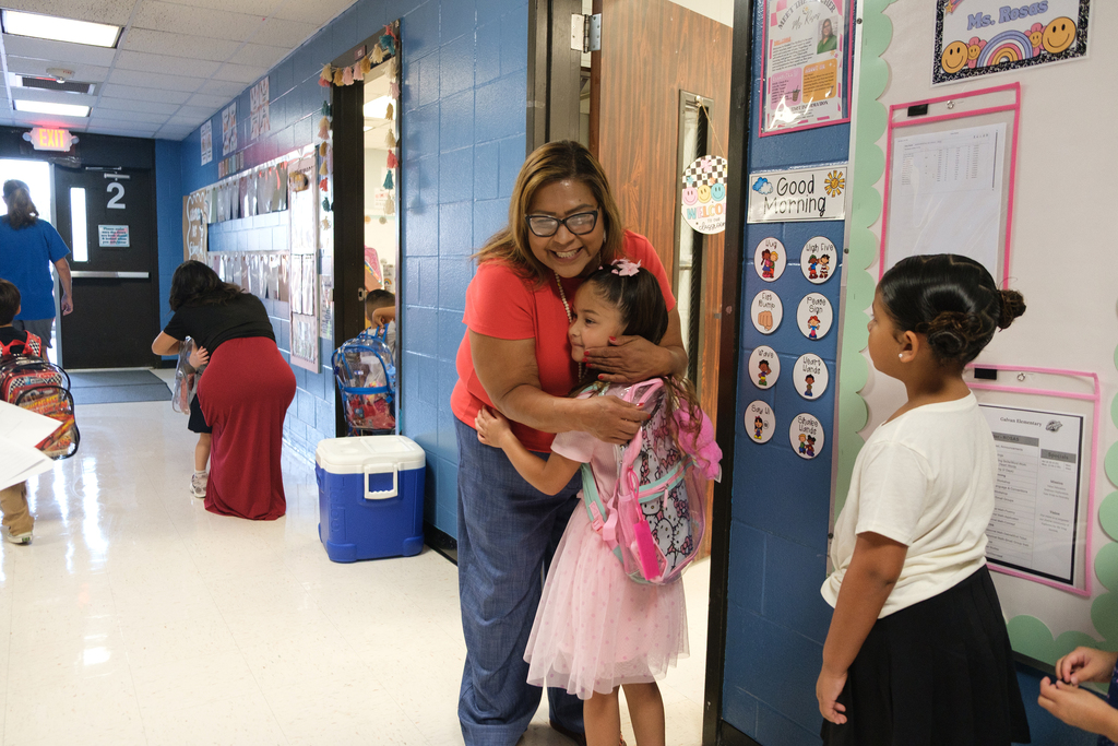 A woman wearing glasses hugs a child in a hallway. Behind them, a girl in a skirt watches.