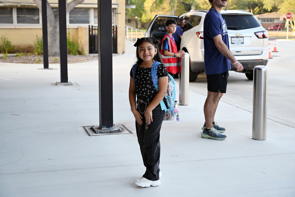 Girl in a black shirt and pants stands in a school parking lot. She wears a backpack.