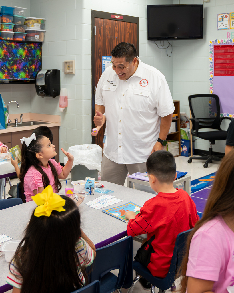 A teacher with a white shirt and a red logo speaks to children around a table in a classroom.