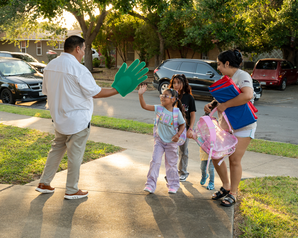 A man in a white shirt and green glove waves at a girl with a backpack, with other children and adults nearby.
