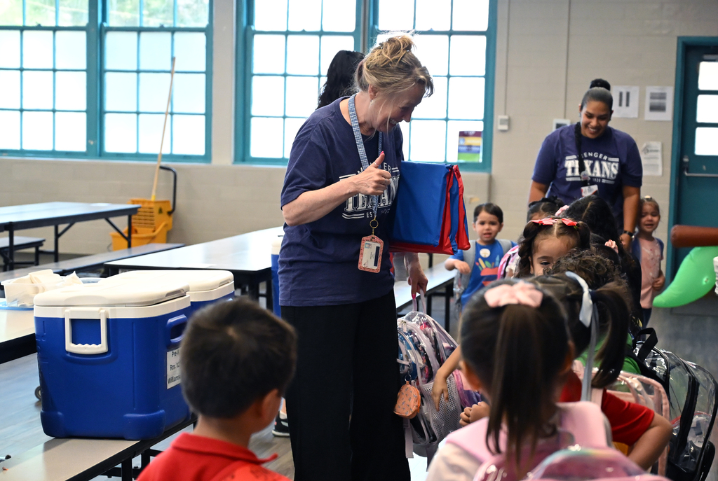 A woman stands with a blue bag in a school cafeteria. Children sit in chairs, some with backpacks. Tables have coolers.