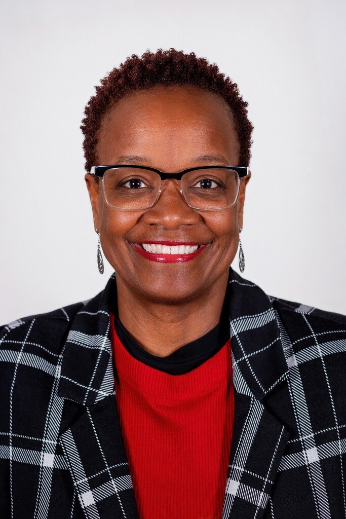 A woman with short hair and glasses smiles. She wears a red shirt under a black and white checkered blazer.
