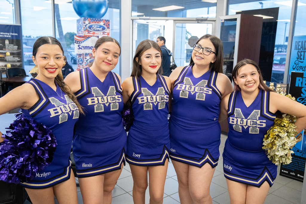 Five cheerleaders in blue uniforms stand close together, smiling, with pom-poms and a blue balloon in the background.