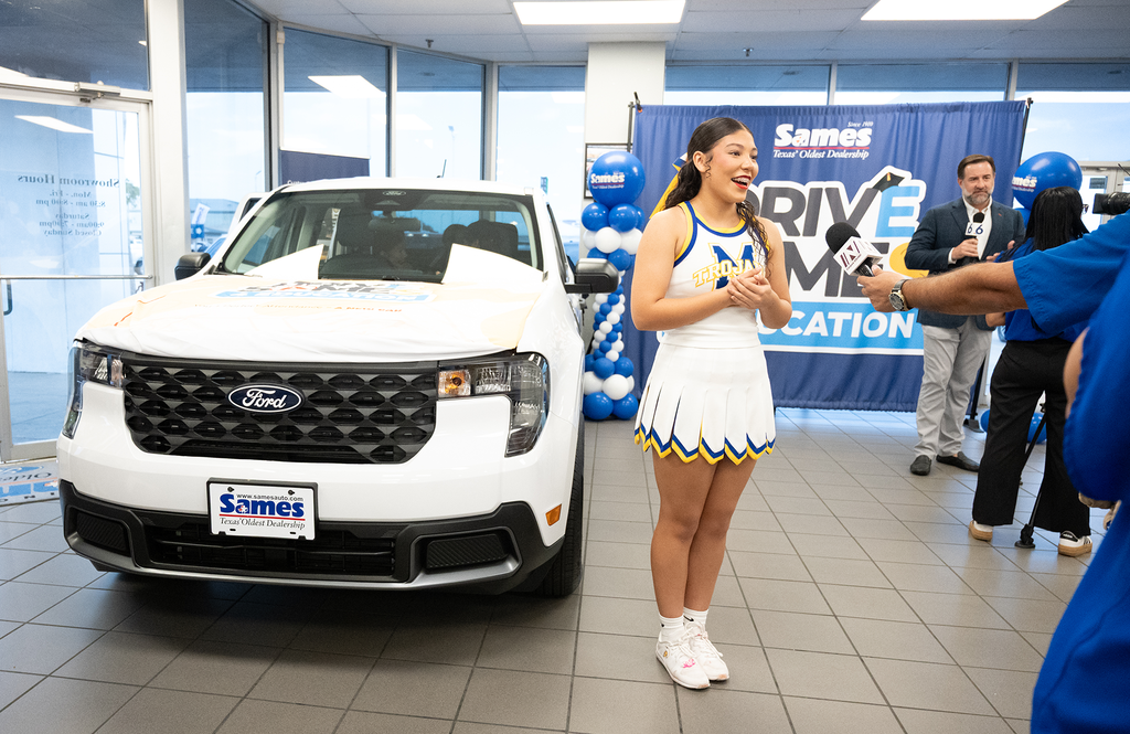 A person stands in front of a white vehicle in a showroom, with a cheerleader in the foreground.