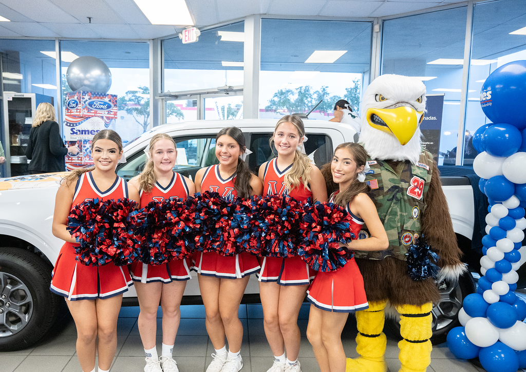 Five cheerleaders in red and white uniforms pose with an eagle mascot in a showroom. Behind them, a white car and blue and white balloons.